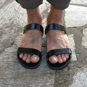 Pode incluir: Sand&aacute;lias de couro preto com um design simples. As sand&aacute;lias t&ecirc;m uma tira larga sobre o p&eacute; e outra ao redor do tornozelo. As solas escuras repousam sobre uma superf&iacute;cie de pedra.