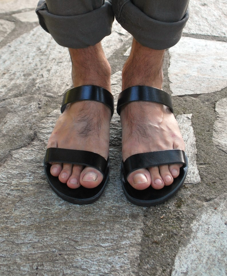 Pode incluir: Sand&aacute;lias de couro preto com um design simples. As sand&aacute;lias t&ecirc;m uma tira larga sobre o p&eacute; e outra ao redor do tornozelo. As solas escuras repousam sobre uma superf&iacute;cie de pedra.
