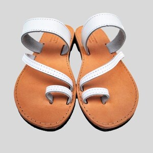 Puede incluir: Un par de sandalias de cuero blanco con presilla para el dedo y correa en el tobillo. Las sandalias tienen una suela marr&oacute;n claro y est&aacute;n dise&ntilde;adas para uso casual. Las sandalias se muestran sobre un fondo gris claro.