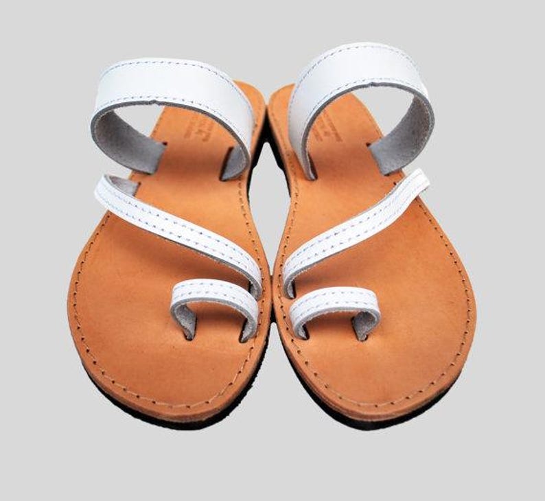 Puede incluir: Un par de sandalias de cuero blanco con presilla para el dedo y correa en el tobillo. Las sandalias tienen una suela marr&oacute;n claro y est&aacute;n dise&ntilde;adas para uso casual. Las sandalias se muestran sobre un fondo gris claro.