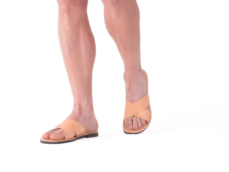 K&ouml;nnte beinhalten: Hellbraune Ledersandalen mit gekreuzten Riemen. Die Sandalen haben eine flache Sohle und werden an den F&uuml;&szlig;en einer Person getragen. Das Bild ist vor einem wei&szlig;en Hintergrund aufgenommen, um die Schuhe hervorzuheben.