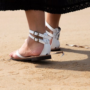 May include: White leather sandals with ankle straps and buckle closures. The sandals have a flat sole and are worn on a sandy beach. The straps are secured with small, antique-style buckles. The sandals are a summer footwear.