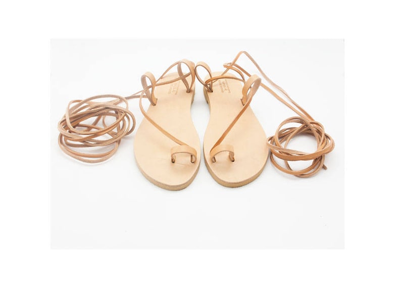 K&ouml;nnte beinhalten: Ein Paar hellbraune Ledersandalen mit Zehenschlaufen und langen, umlaufenden Kn&ouml;chelriemen. Die Sandalen haben ein schlichtes Design mit flacher Sohle und werden auf wei&szlig;em Hintergrund pr&auml;sentiert. Die Riemen sind zur Seite gerollt.