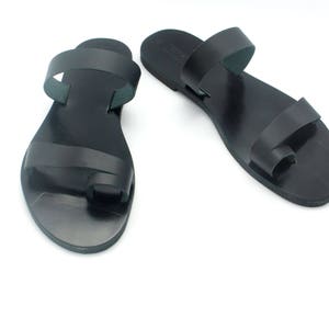 May include: A pair of black leather sandals with a toe loop and two straps. The sandals are flat and have a simple, minimalist design. The sandals are shown on a white background.