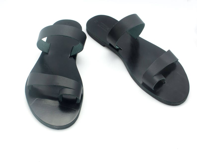 May include: A pair of black leather sandals with a toe loop and two straps. The sandals are flat and have a simple, minimalist design. The sandals are shown on a white background.