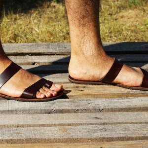 May include: Brown leather sandals with two straps across the foot and a toe loop. The sandals are worn on a wooden surface, with a grassy background visible. The sandals appear to be handmade.