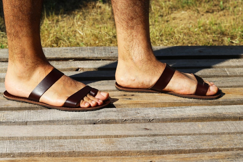May include: Brown leather sandals with two straps across the foot and a toe loop. The sandals are worn on a wooden surface, with a grassy background visible. The sandals appear to be handmade.