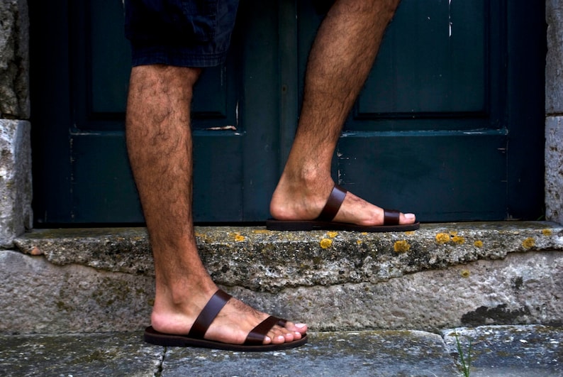 May include: Close-up of brown leather sandals with two straps. The sandals are worn on a person's feet, with the legs visible. The person is standing on a stone step in front of a dark green door.