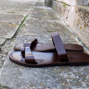 May include: A pair of brown leather sandals with a simple design. The sandals have a flat sole and two straps across the top of the foot. The sandals are made of a smooth, shiny leather and are sitting on a stone surface.