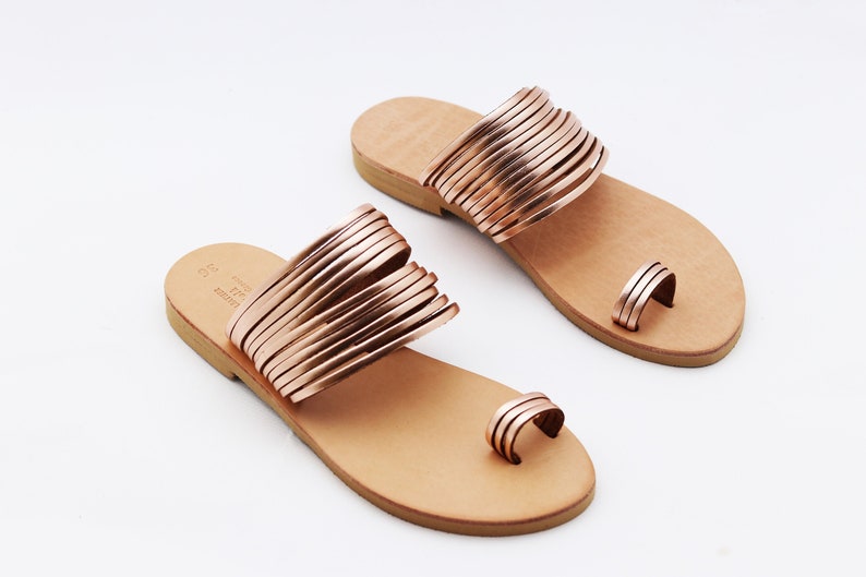 May include: A pair of rose gold sandals with a toe loop and multiple straps across the top. The sandals have a light brown leather footbed and a simple design, perfect for warm weather. The sandals are displayed on a white background.