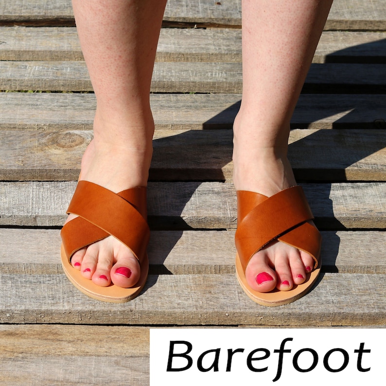 Peut inclure: Sandales en cuir marron avec un design crois&eacute;, pr&eacute;sent&eacute;es sur une surface en bois. Le mot "Barefoot" est &eacute;crit en blanc au bas de l'image. Les pieds sont visibles.