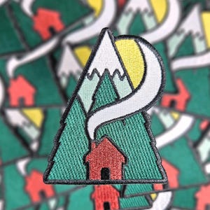 May include: Embroidered patch depicting a red cabin in a green forest, with mountains and a yellow sun. White smoke curls from the cabin's chimney. The patch has a black border. Ideal for jackets or bags.