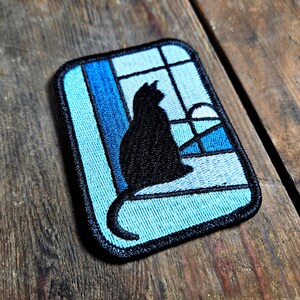 May include: Embroidered patch depicting a black cat silhouette in a window. The window design uses shades of blue, with a black border. The patch is rectangular and has a black border.