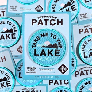 May include: Embroidered patch with the text "TAKE ME TO THE LAKE" in black on a light blue background. The patch features a mountain and sun design. The patch is surrounded by a light blue border. The words "IRON-ON OR SEW YOU CHOOSE!" are also visible.