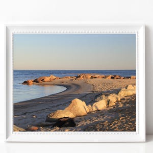 Puede incluir: Fotografía enmarcada de una escena costera. La imagen presenta una playa de arena con un muelle de roca curvado que se extiende hacia el océano. El cielo es un degradado de azul y el agua está en calma.