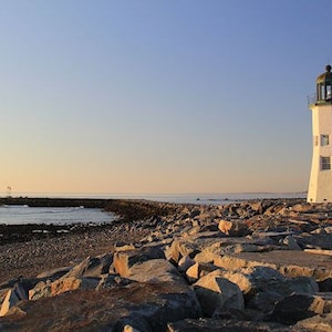 May include: A white lighthouse with a green roof stands on a rocky shore. The lighthouse is surrounded by large rocks and the ocean is in the background. The sky is a light blue color.