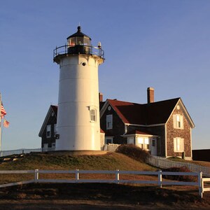 May include: A white lighthouse with a black roof and a red brick house with a red roof. The lighthouse is on a grassy hill with a white picket fence in front of it. The sky is blue and there is a small American flag flying in the wind.