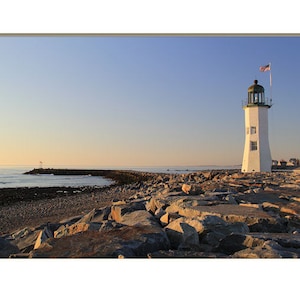 May include: A white lighthouse with a green roof stands on a rocky shore. The lighthouse is surrounded by rocks and the ocean is in the background. The sky is a light blue color.