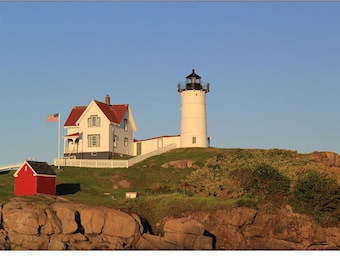 May include: A coastal scene featuring a white lighthouse with a black top, a red-roofed house, and a small red shed. The structures sit atop a grassy hill with a clear blue sky in the background. An American flag waves.