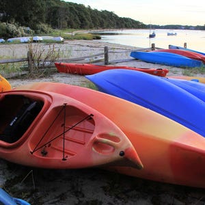 Puede incluir: Una colección de coloridos kayaks, incluyendo rojos, naranjas y azules, descansan en una playa arenosa. Los kayaks son de varios tamaños y diseños, sugiriendo opciones para actividades acuáticas. El fondo muestra un cuerpo de agua tranquilo y una línea de árboles.