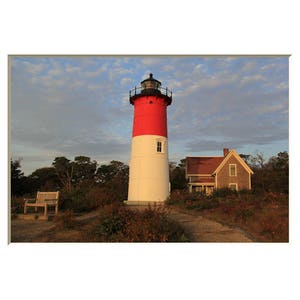 Puede incluir: Un faro rojo y blanco con un techo negro se alza alto contra un cielo azul con nubes blancas. Una pequeña casa marrón con molduras blancas se encuentra a la derecha del faro. Un banco de madera se encuentra en primer plano.
