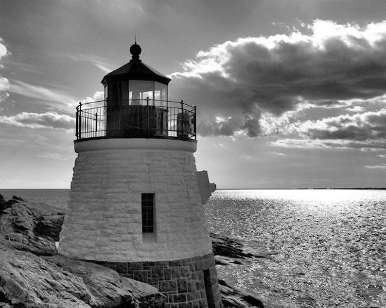 Pu&ograve; includere: Una fotografia in bianco e nero di un faro su una costa rocciosa. Il faro &egrave; alto e bianco con un tetto nero e una ringhiera nera. Il cielo &egrave; nuvoloso e l'acqua &egrave; calma.