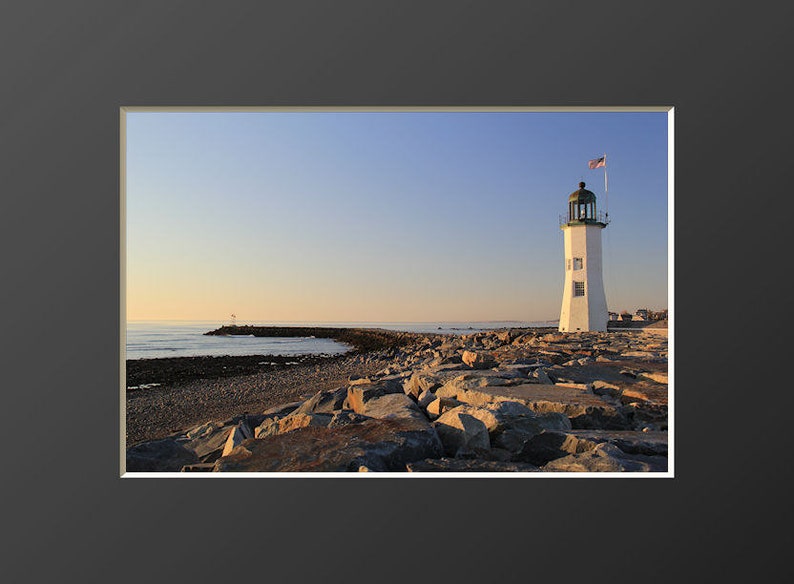May include: A white lighthouse with a green roof stands on a rocky shore at sunset. The lighthouse is tall and slender, with a small flag flying from its top. The rocks are gray and brown, and the water is calm and blue.