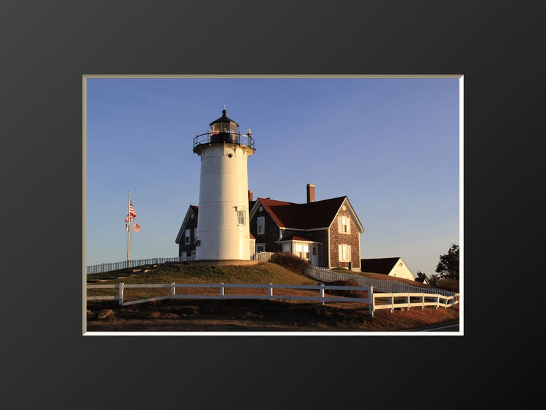 May include: A white lighthouse with a red roof and a black lantern on top, standing on a grassy hill with a white picket fence in front. The lighthouse is surrounded by a blue sky and a few trees in the background.