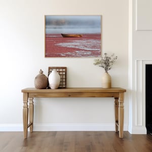Puede incluir: Una mesa consola de madera con un acabado marrón claro. La mesa tiene una tapa rectangular y dos patas torneadas a cada lado. Hay una fotografía enmarcada de un barco en un lago con un fondo rojo colgada en la pared sobre la mesa. Hay dos jarrones decorativos y un marco de madera sobre la mesa.