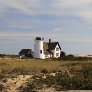 Puede incluir: Un faro blanco con una ventana roja y un techo negro se alza sobre una colina cubierta de hierba. Un edificio blanco más pequeño con un techo marrón se encuentra detrás del faro.