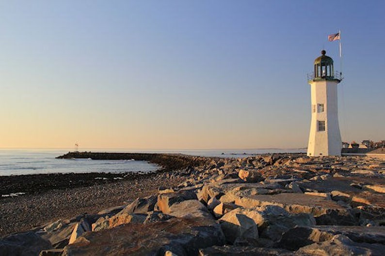 May include: A white lighthouse with a green roof stands on a rocky shore. The lighthouse is surrounded by large rocks and the ocean is in the background. The sky is a light blue color.
