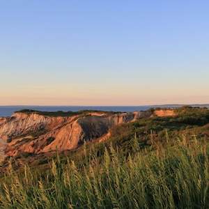 Puede incluir: Un faro de ladrillo rojo se alza sobre un acantilado con vistas a una playa de arena y el océano. El cielo es de un azul pálido con un toque de rosa del sol poniente. Hierba verde y arbustos cubren la ladera del acantilado.