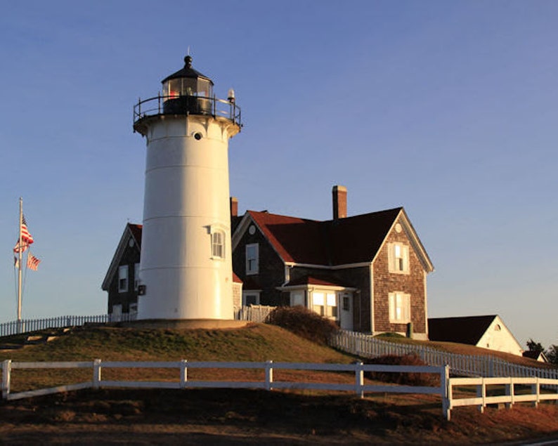 May include: A white lighthouse with a black roof and a red brick house with a red roof. The lighthouse is on a grassy hill with a white picket fence in front of it. The sky is blue and there is a small American flag flying in the wind.