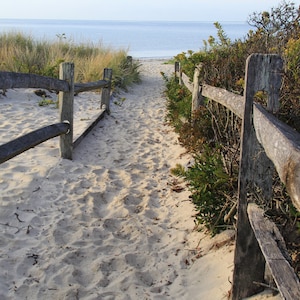 Puede incluir: Una valla de madera conduce a un camino de arena que termina en una playa con agua azul en la distancia.
