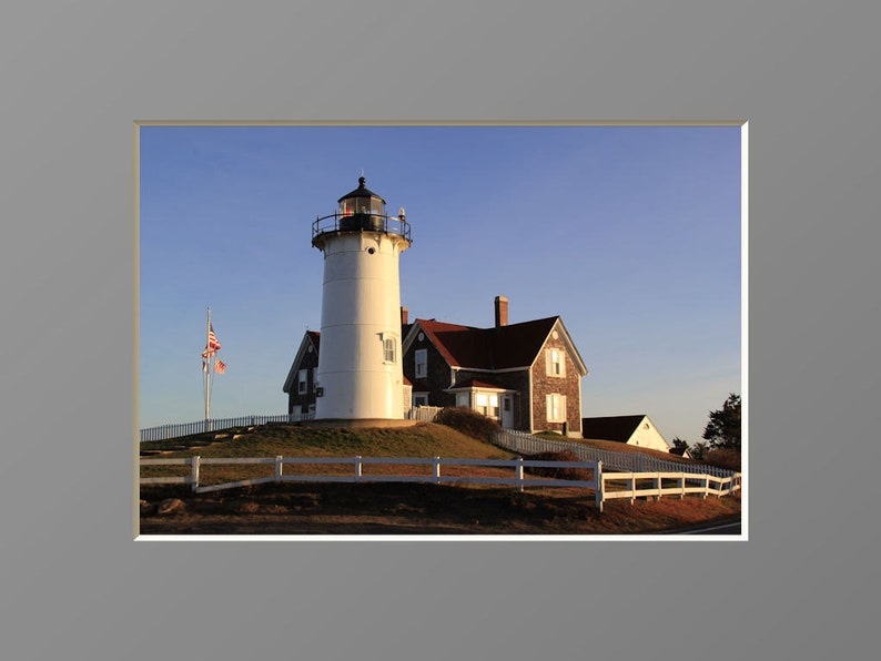May include: A white lighthouse with a red roof and a black lantern on top. The lighthouse is on a grassy hill with a white picket fence in front of it. There is an American flag flying in the wind.