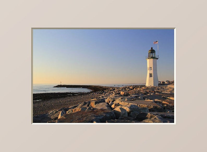 May include: A white lighthouse with a green roof stands on a rocky shore. The lighthouse is surrounded by rocks and the ocean is in the background. The sky is a light blue color.
