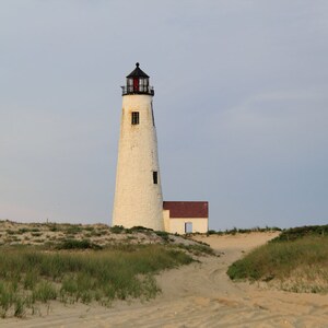 Puede incluir: Un faro blanco con techo negro y puerta roja se alza en una playa de arena con hierba verde. El faro es alto y delgado, con un pequeño edificio adjunto.