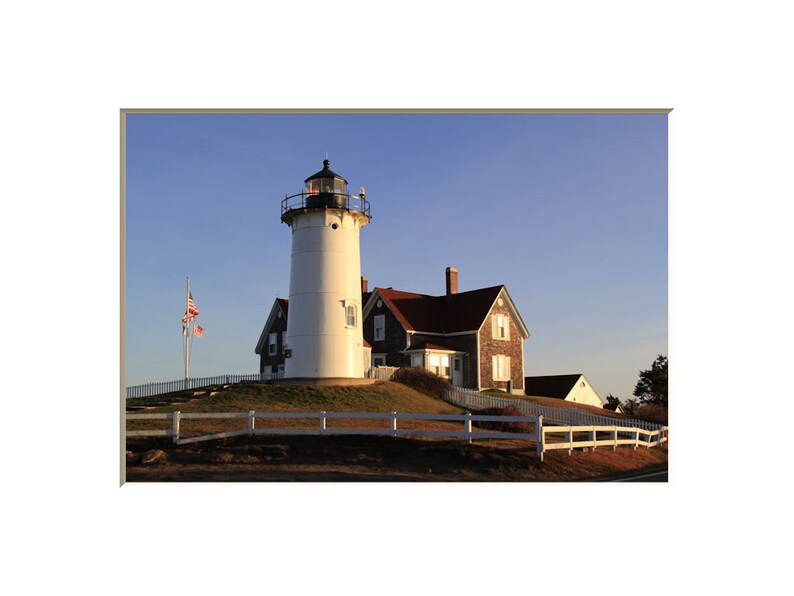 May include: A white lighthouse with a red roof and a black lantern on top. The lighthouse is on a grassy hill with a white picket fence in front of it. An American flag is flying on a pole in front of the lighthouse.