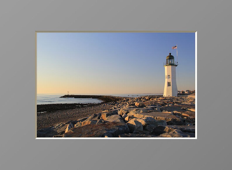 May include: A white lighthouse with a green roof stands on a rocky outcropping. The lighthouse is on a small island with a body of water in the background. The sky is a light blue color.