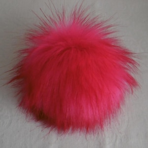 Könnte beinhalten: Ein leuchtend pinkfarbener Kunstfell-Bommel. Der runde Bommel hat lange, flauschige Strähnen, die nach außen strahlen. Die leuchtende Farbe und Textur machen ihn zu einem auffälligen Accessoire für Hüte, Schals oder andere Bastelarbeiten.