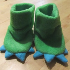May include: A pair of green fleece baby booties with blue felt dinosaur claws.