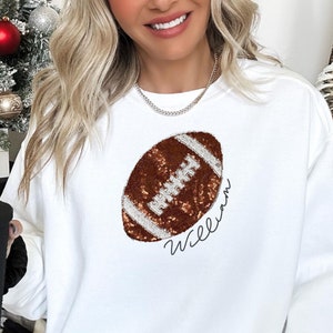 Könnte beinhalten: Weißer Pullover mit einem braunen und weißen Pailletten-Football-Design und dem Namen "William" in Kursivschrift.