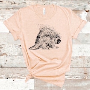 K&ouml;nnte beinhalten: Ein helles, pfirsichfarbenes T-Shirt mit einer Schwarz-Wei&szlig;-Zeichnung eines Stachelschweins auf der Vorderseite.