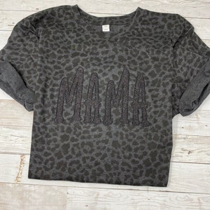 May include: A dark grey sweatshirt featuring a leopard print design. The word "MAMA" is embroidered across the front in a glittery, dark grey thread. The sleeves are cuffed, and the shirt has a crew neck.