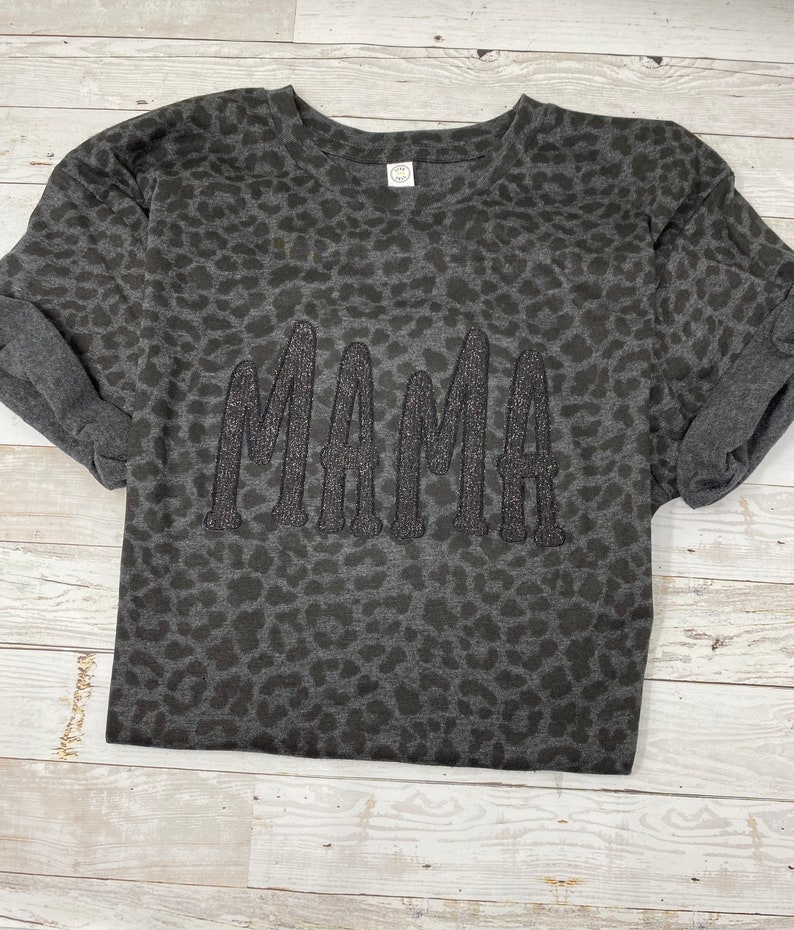 May include: A dark grey sweatshirt featuring a leopard print design. The word "MAMA" is embroidered across the front in a glittery, dark grey thread. The sleeves are cuffed, and the shirt has a crew neck.