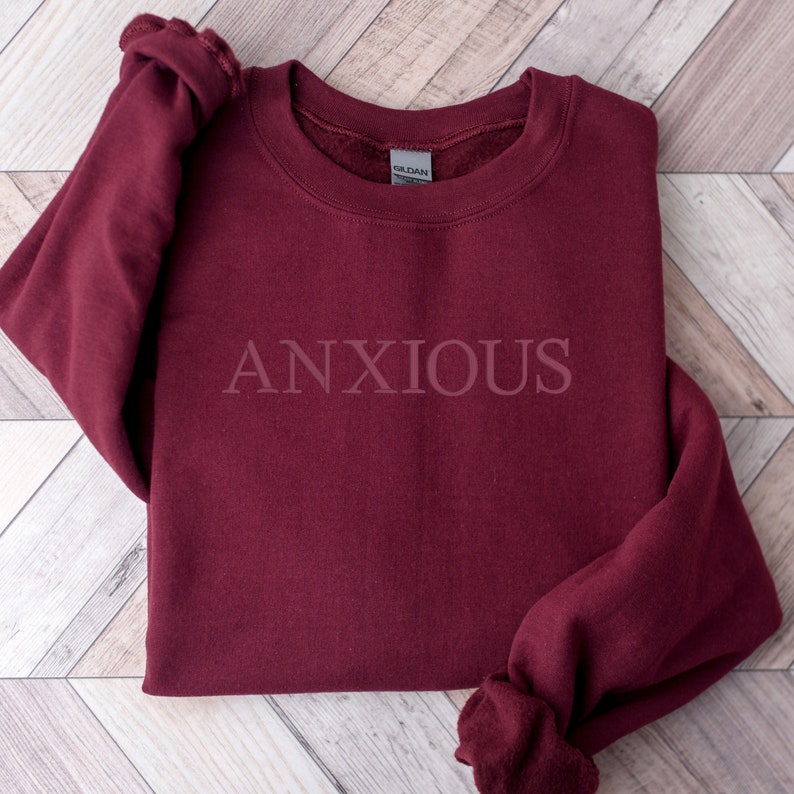 May include: Burgundy sweatshirt with the word "ANXIOUS" printed in white on the front.