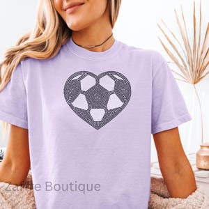 Peut inclure: Un t-shirt lavande avec un motif de ballon de football en strass noir en forme de cœur. Le t-shirt a un col rond et des manches courtes.