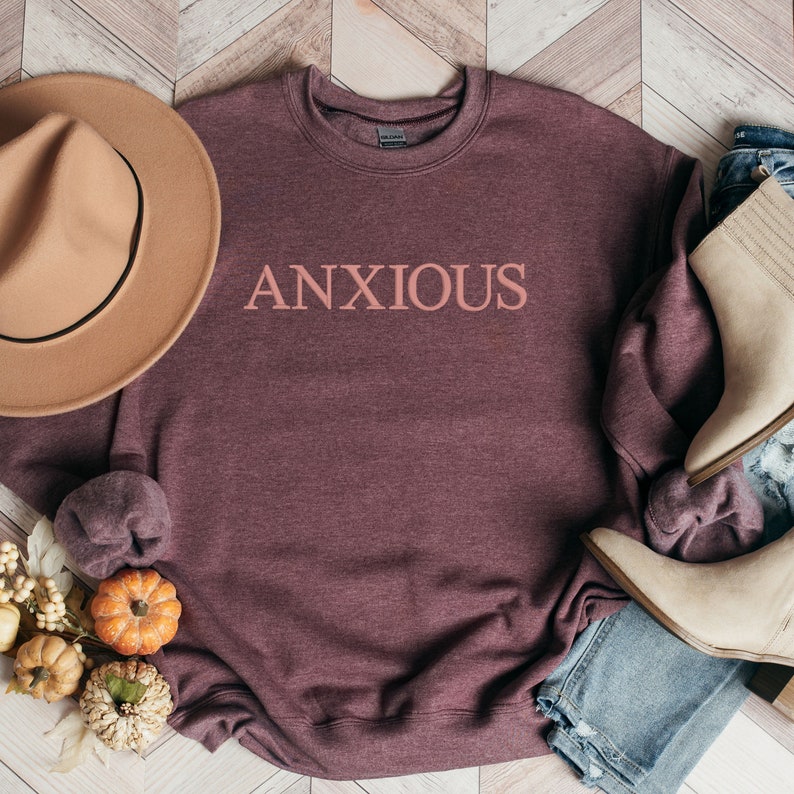 May include: A maroon sweatshirt with the word "ANXIOUS" printed in pink on the front. The sweatshirt is laid flat on a wooden surface with a brown hat, jeans, and boots.