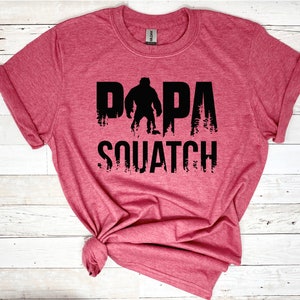 May include: A red t-shirt with a black graphic design that says "Papa Squatch" with a silhouette of Bigfoot in the middle of the word "Papa".