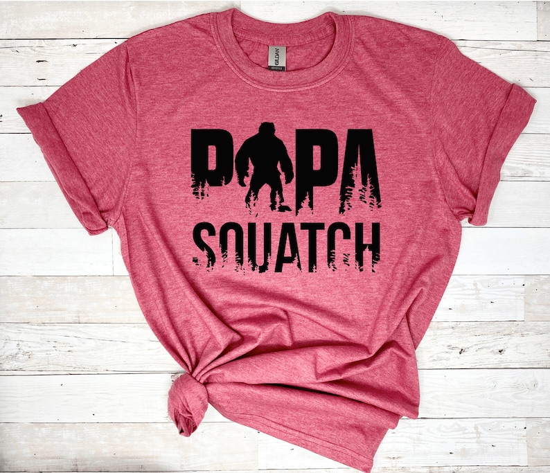 May include: A red t-shirt with a black graphic design that says "Papa Squatch" with a silhouette of Bigfoot in the middle of the word "Papa".